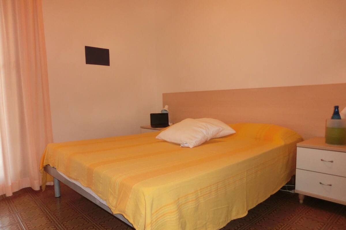 Un lit simple avec une couvre-lit jaune, un oreiller blanc, une table de chevet avec une lampe et une bouteille d’eau sur la table d’appoint.