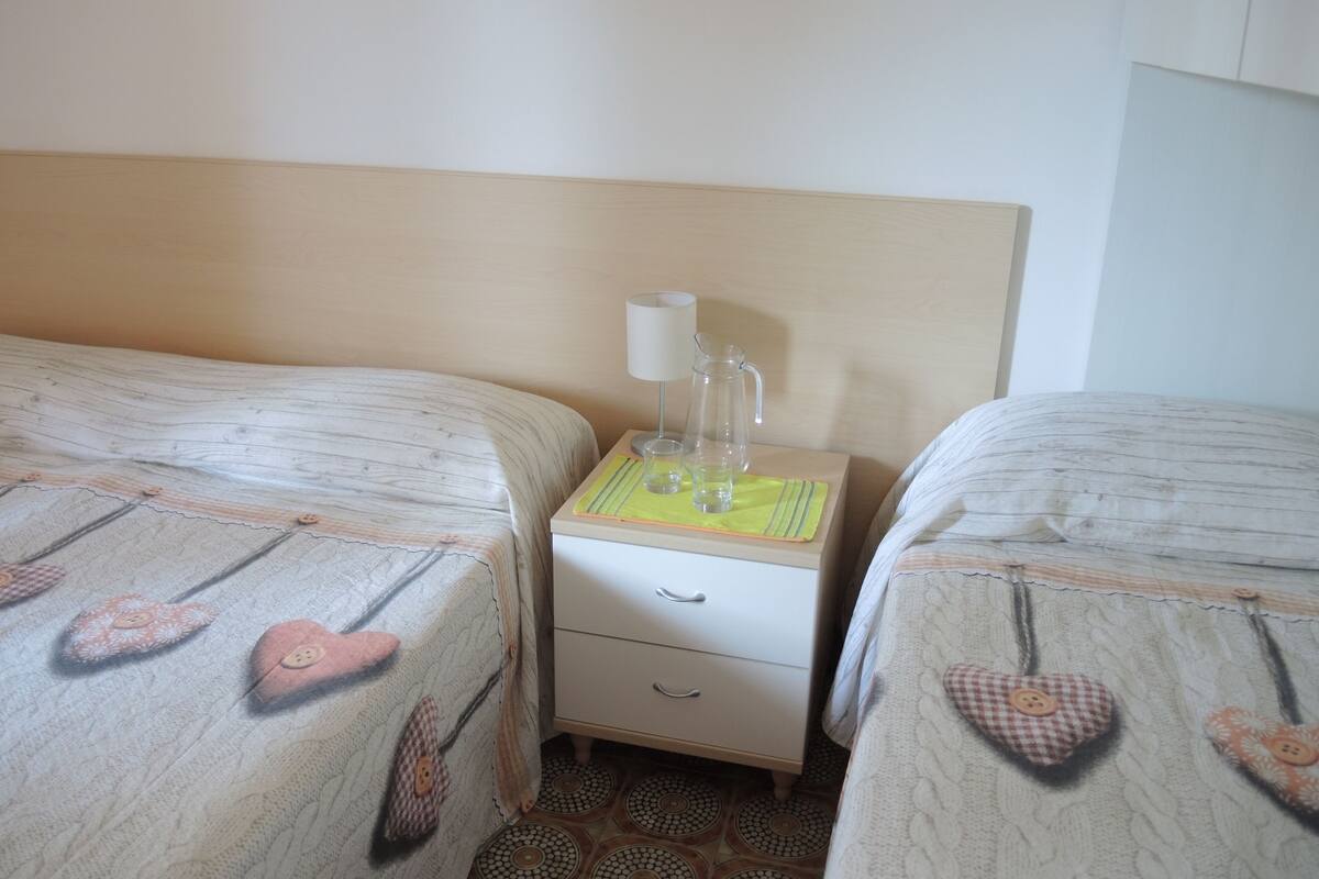 Une chambre d’hôtel avec deux lits, une table de chevet avec une lampe, une carafe et un petit tiroir.