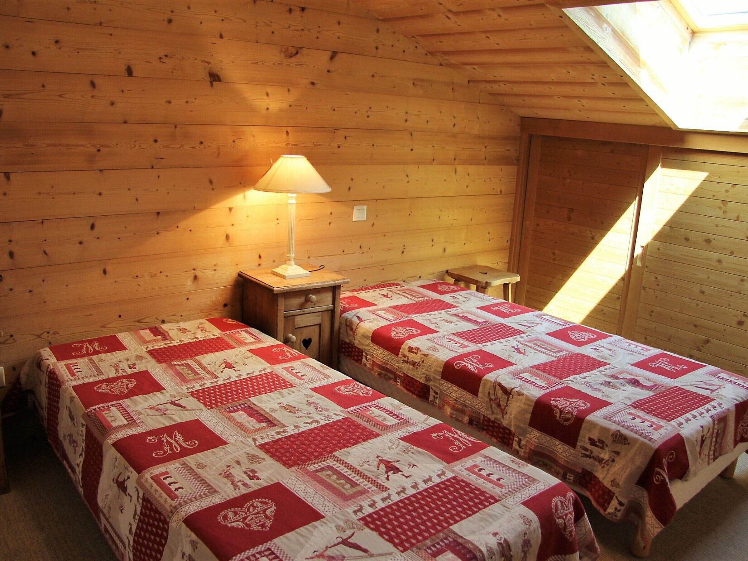 Une pièce chaleureuse avec deux lits, chacun recouvert d’une couverture rouge et blanche, une petite table de chevet en bois avec une lampe, et un plafond en pente doté d’un puits de lumière.