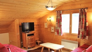 Acogedora habitación tipo cabaña de madera con paredes de madera, un televisor, una mesita, dos sofás rojos y una ventana con cortinas.