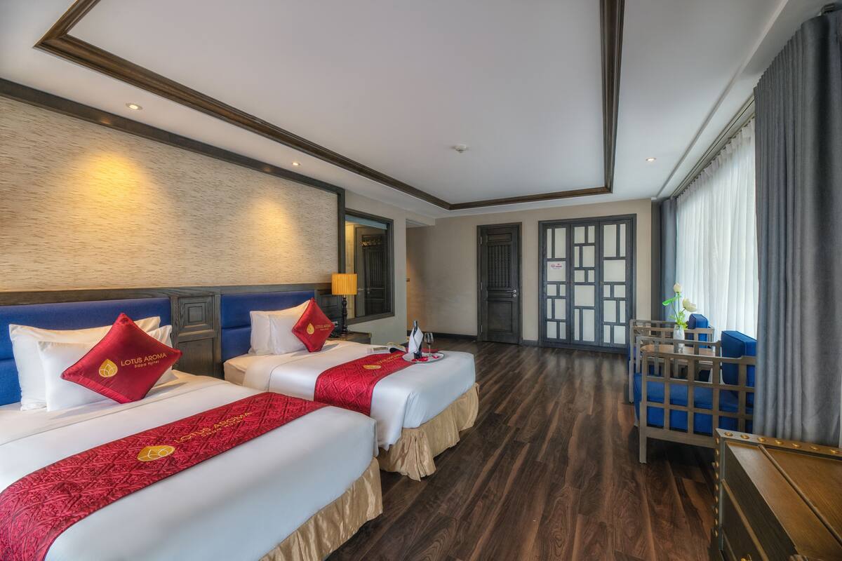 Habitación de hotel con dos camas, piso de madera, un ventanal grande con cortinas y una mesita con un jarrón.