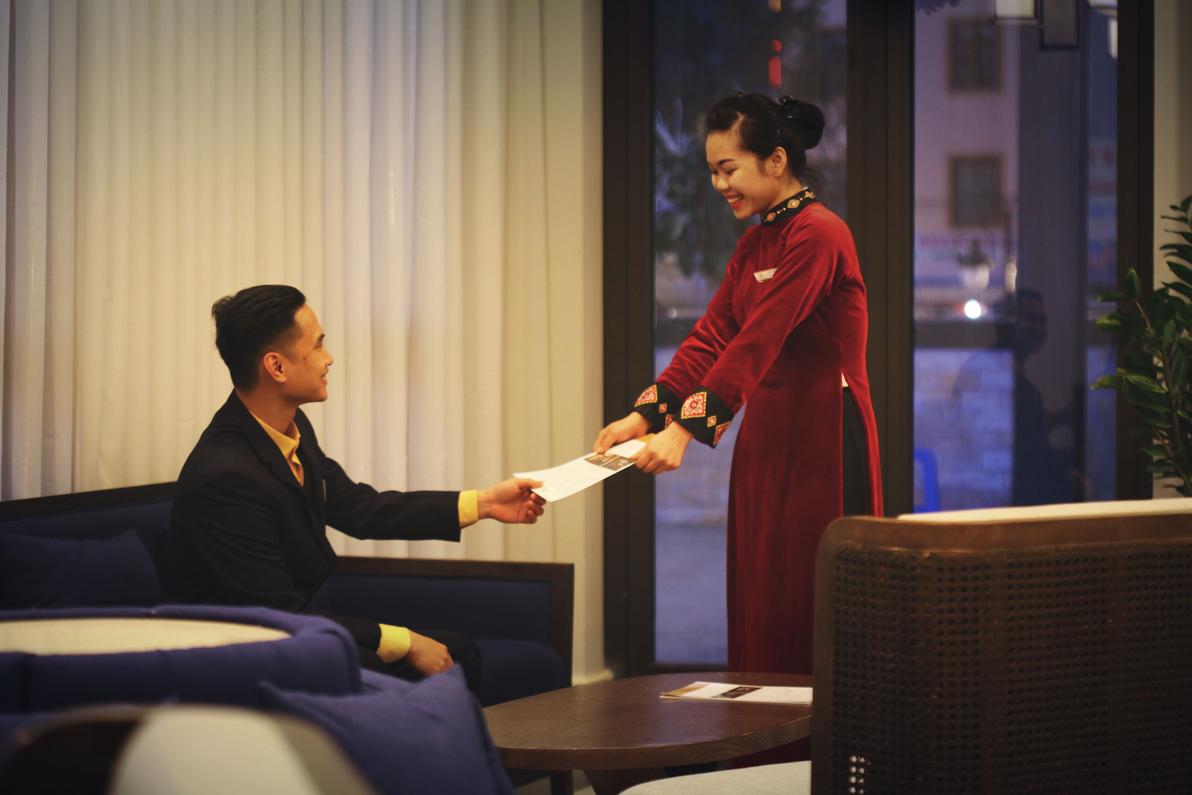 A man and a woman in a hotel lobby, exchanging a document.
