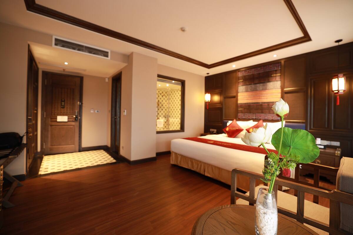 Una habitación de hotel con piso de madera, una cama con ropa de cama roja y blanca, un jarrón con flores y una silla de madera.