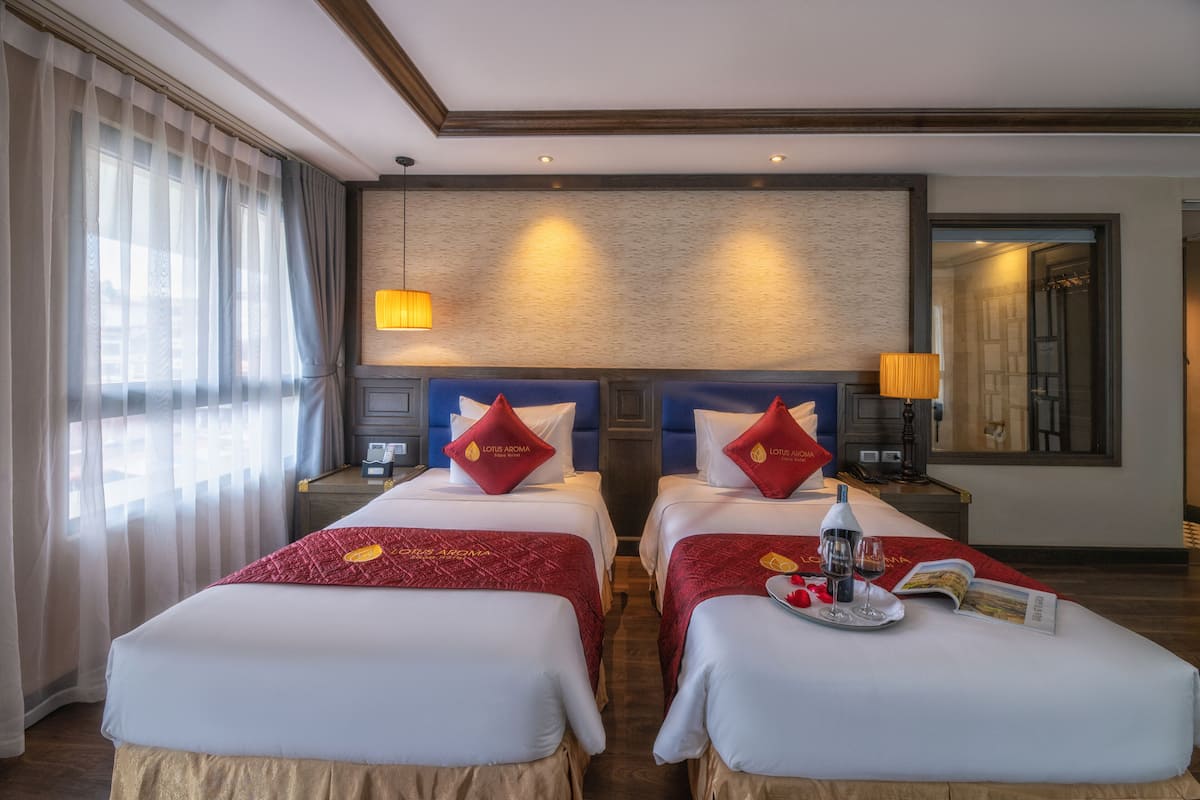 Habitación de hotel con dos camas, cada una con un almohadón rojo, una mesita de noche con lámpara y una bandeja con vasos y una revista.