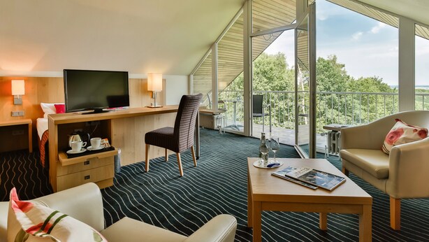 A hotel room with a large window, a television, a desk, a chair, a sofa, and a coffee table. - Berghotel Hohe Mark (Reken)