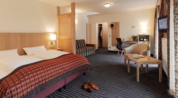 Una habitación de hotel con una cama grande, un escritorio con una silla, una mesita con una taza y un libro, y un televisor en la pared.