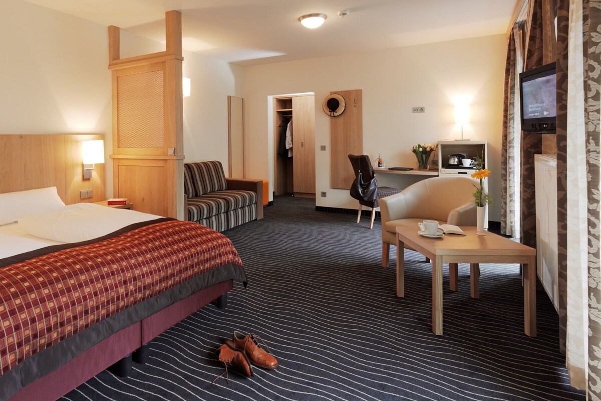 A hotel room with a large bed, a desk with a chair, a small table with a cup and book, and a TV on the wall.