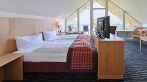 A hotel room with a large bed, a desk with a television, and a view of the outdoors through large windows. - Berghotel Hohe Mark (Reken)