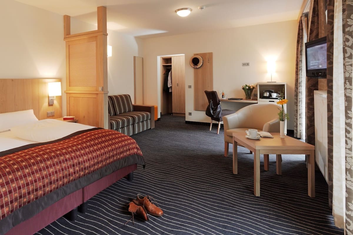 A hotel room with a large bed, a desk with a chair, a small table with a cup and book, and a TV on the wall.