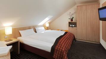 Ein Hotelzimmer mit einem großen Bett, einem Schreibtisch mit Lampe, einem an der Wand befestigten Fernseher und einem Kleiderschrank.