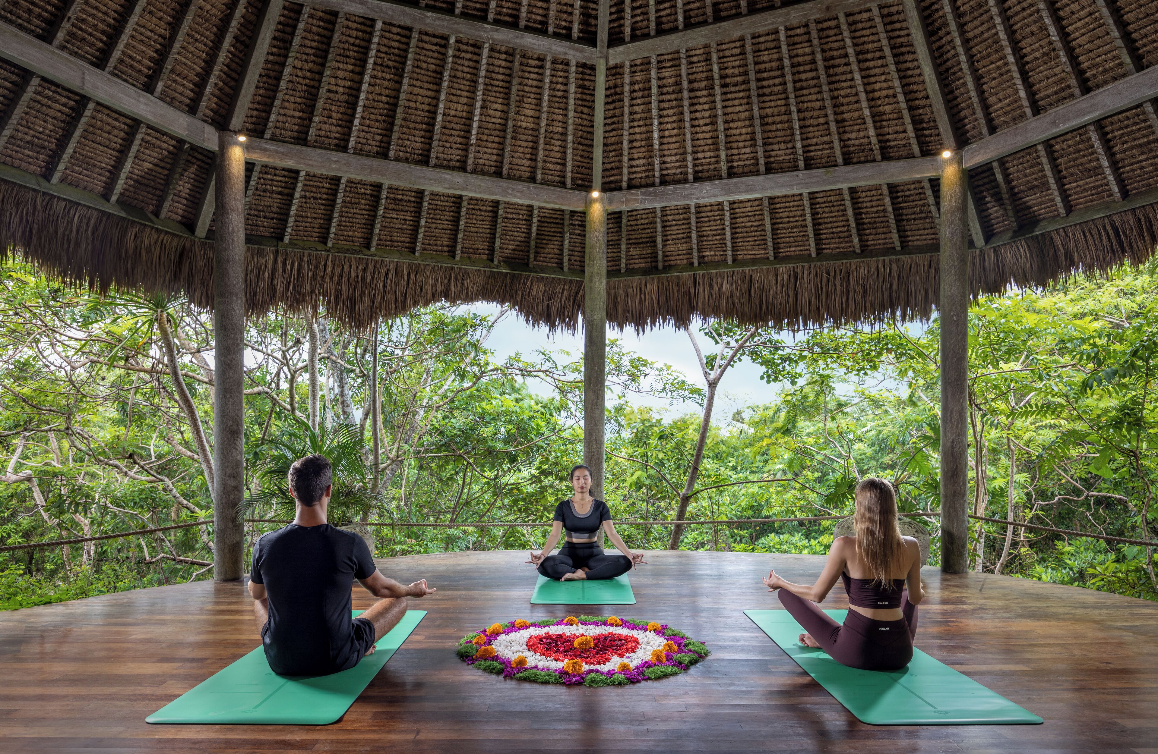 Trois personnes méditent sur des tapis verts dans un pavillon en bois doté d’un toit de chaume, entouré d’une végétation luxuriante.