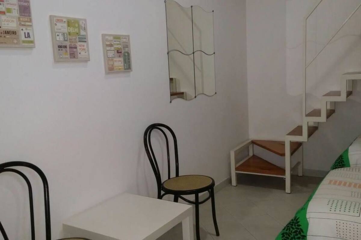 A room with a white wall, a black metal chair, a white table, a bed with a green and white patterned bedspread, and a staircase with a white railing.