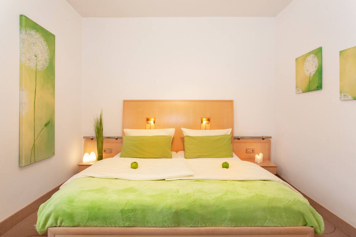 Un lit bien fait, avec une literie verte et deux pommes posées sur les oreillers, de part et d’autre de tables de chevet ornées de bougies et de décorations murales.