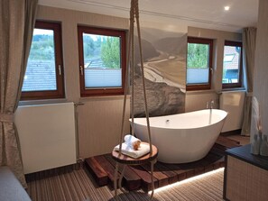 Un baño moderno con una bañera independiente, una plataforma de madera con una toalla y vistas a un río a través de amplias ventanas.