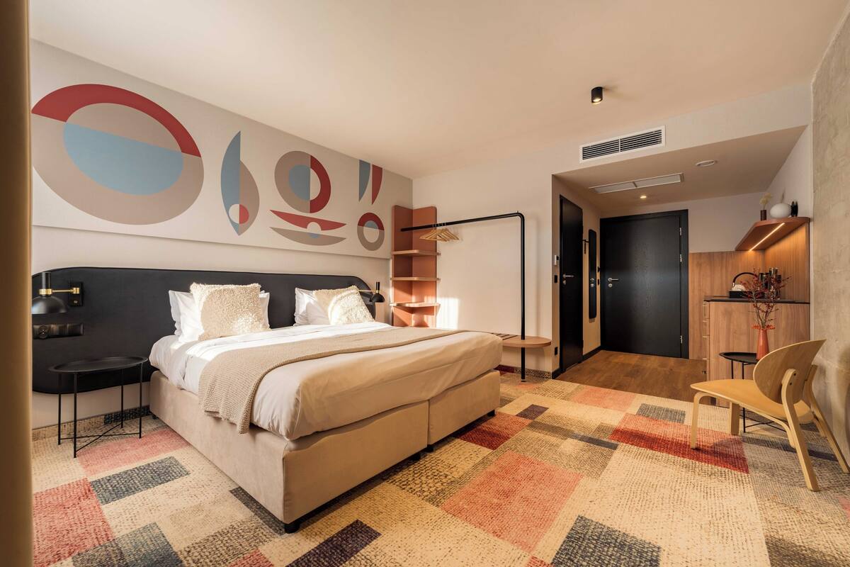 A modern bedroom with a large bed, a nightstand, a chair, a desk, and a wall with abstract art.