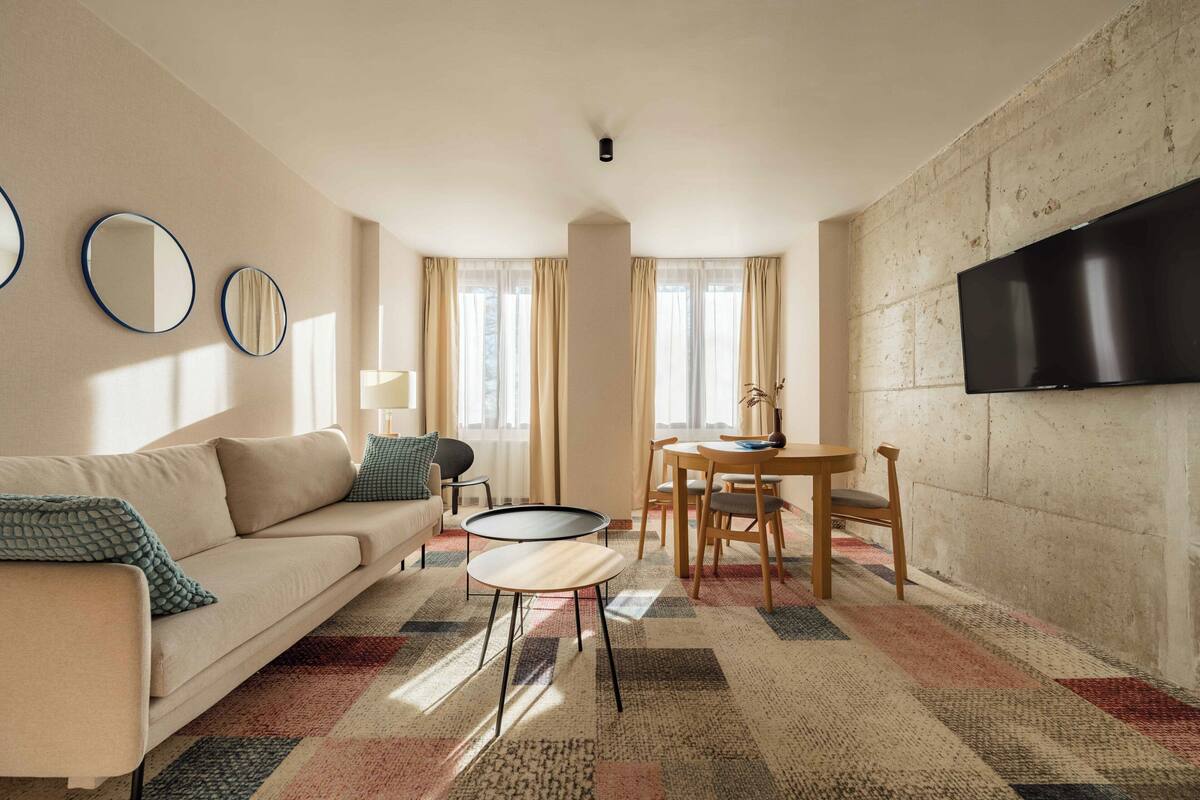 A living room with a sofa, a coffee table, a TV mounted on the wall, and a dining area with chairs and a table.