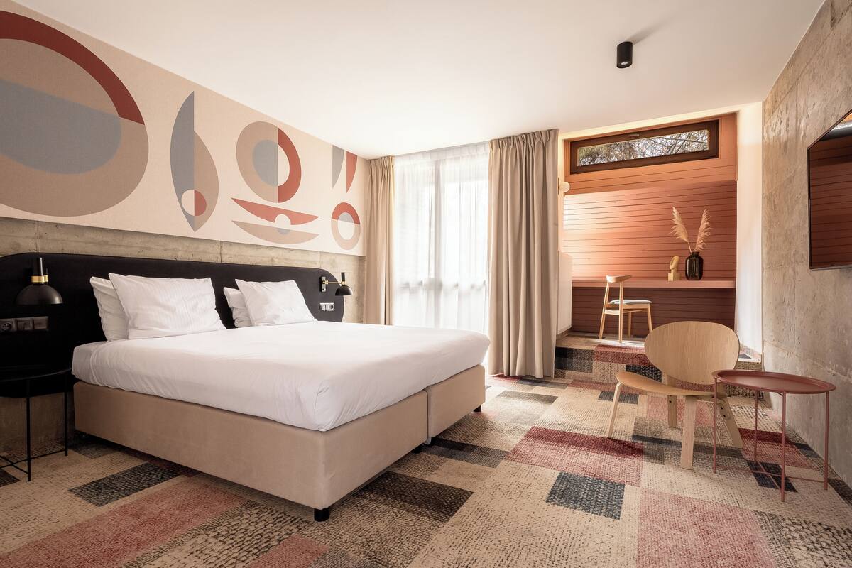 A modern hotel room with a large bed, a desk, and a chair. There is a window with curtains, a wall with abstract art, and a small table with a vase.