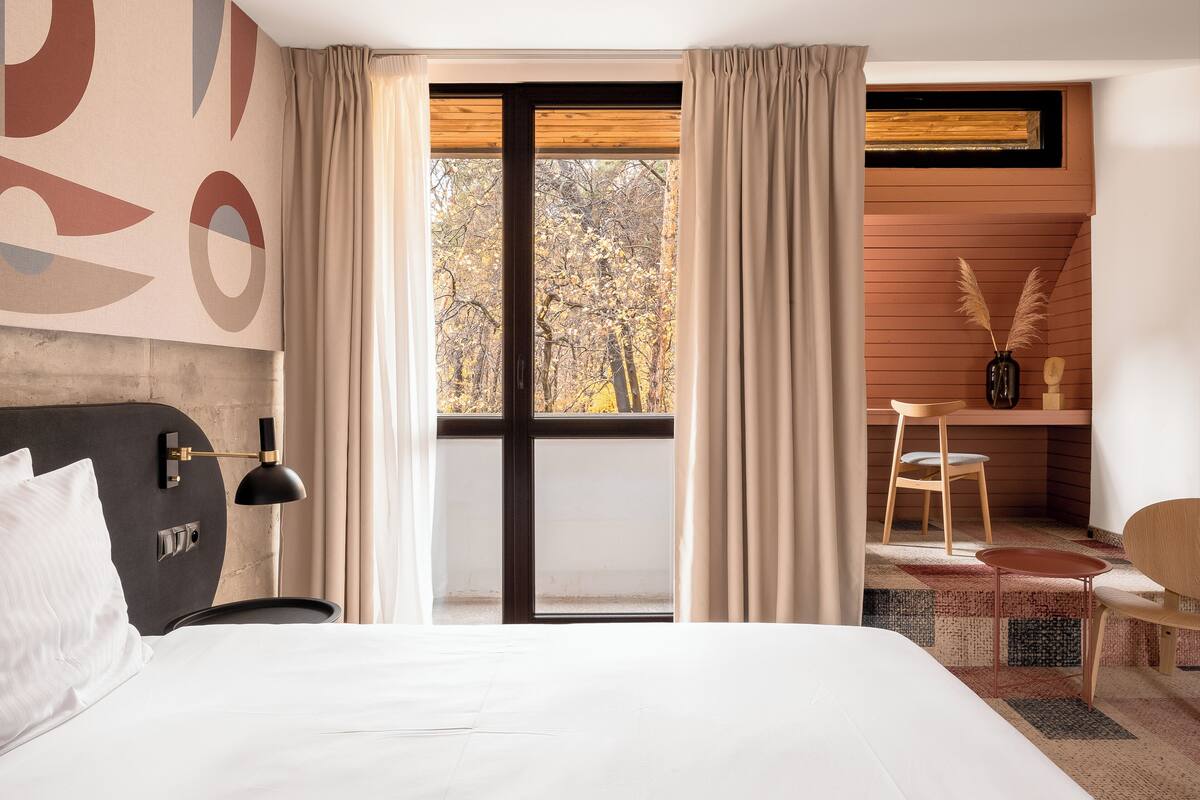 A modern hotel room with a large bed, a desk with a chair, a small table, and a view of trees through the window.