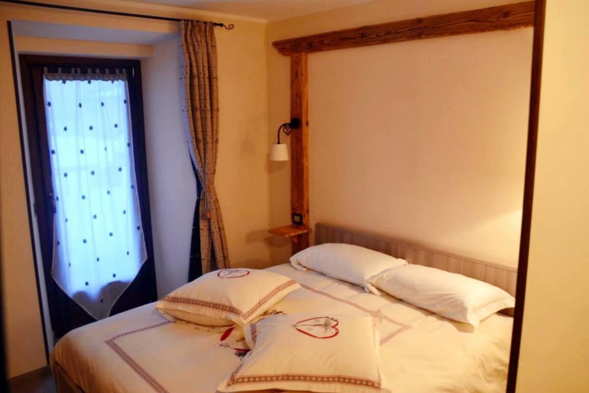 Une chambre à coucher avec un lit, des oreillers, une table de chevet, une lampe et une fenêtre avec des rideaux.