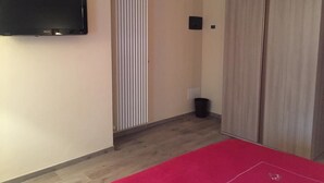 Ein Zimmer mit flachbildfernseher an der Wand, einer Schiebetür zum Kleiderschrank, einem roten Teppich und einer kleinen schwarzen Mülltonne.