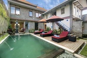 En swimmingpool med liggestole og parasoller, et stenhus med trappe og en have.