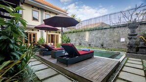 Un área de piscina con sillones de descanso y una sombrilla, rodeada de muros de piedra y vegetación.