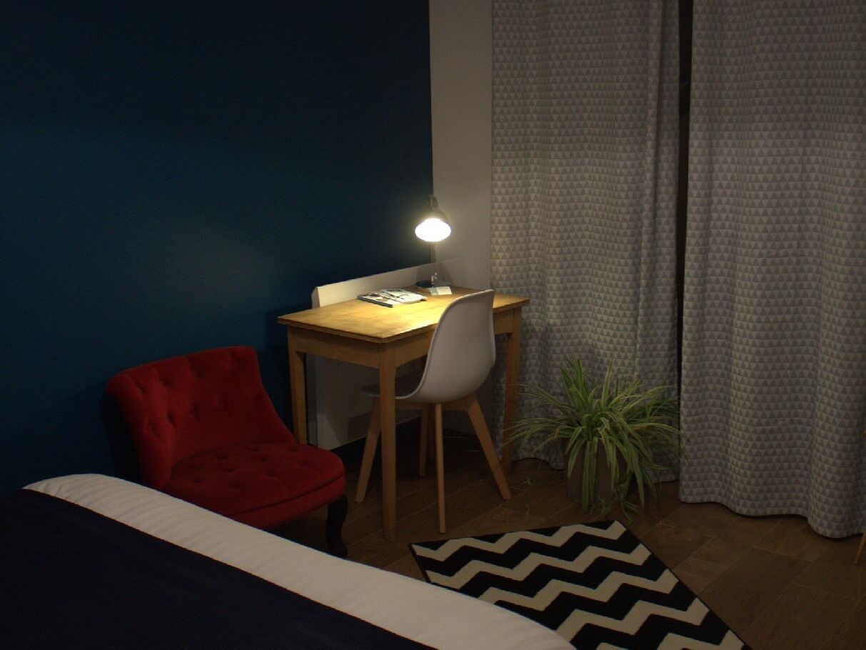 Een kleine kamer met een bed, een bureau, een rode stoel, een lamp en een potplant.
