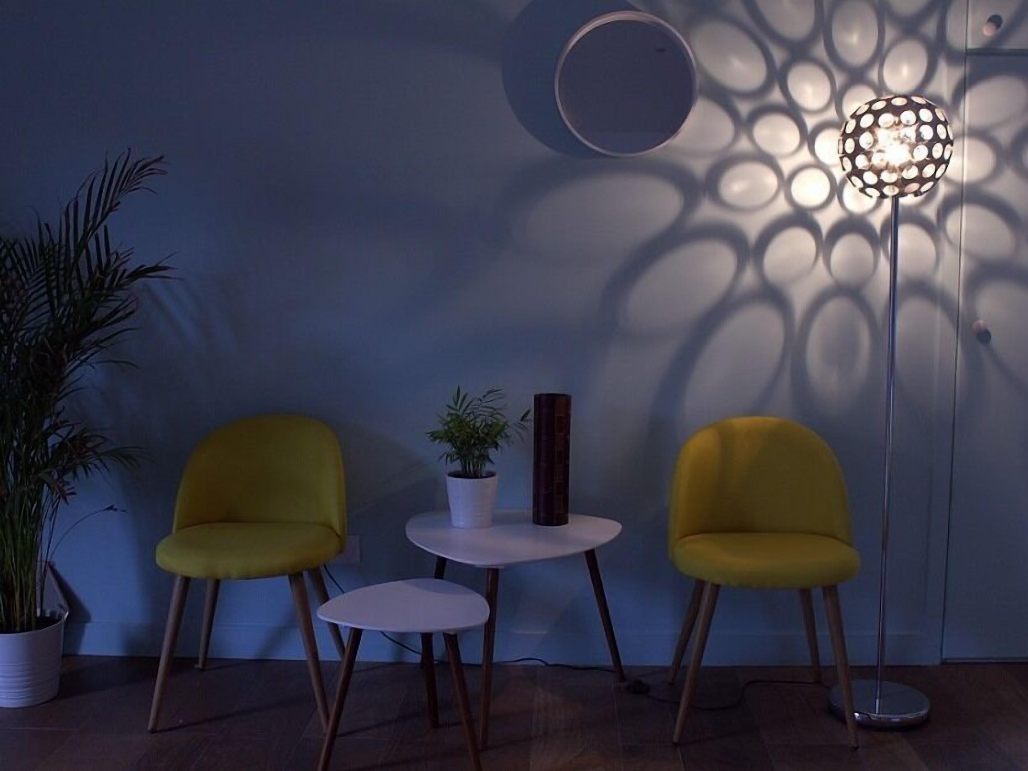 A room with two yellow chairs, a small white table, a potted plant, and a floor lamp casting patterned shadows on the wall.