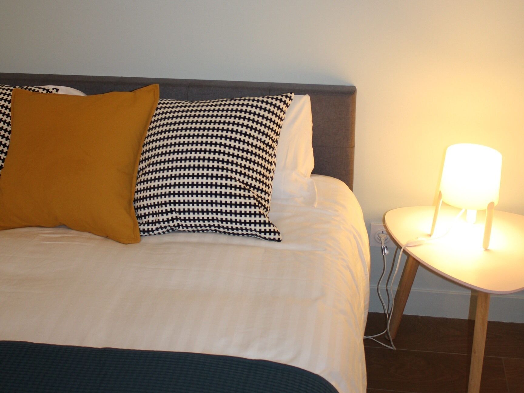 Un lit bien fait, avec une couette blanche et deux coussins à motifs, une petite table de chevet avec une lampe, et un mur sobre.