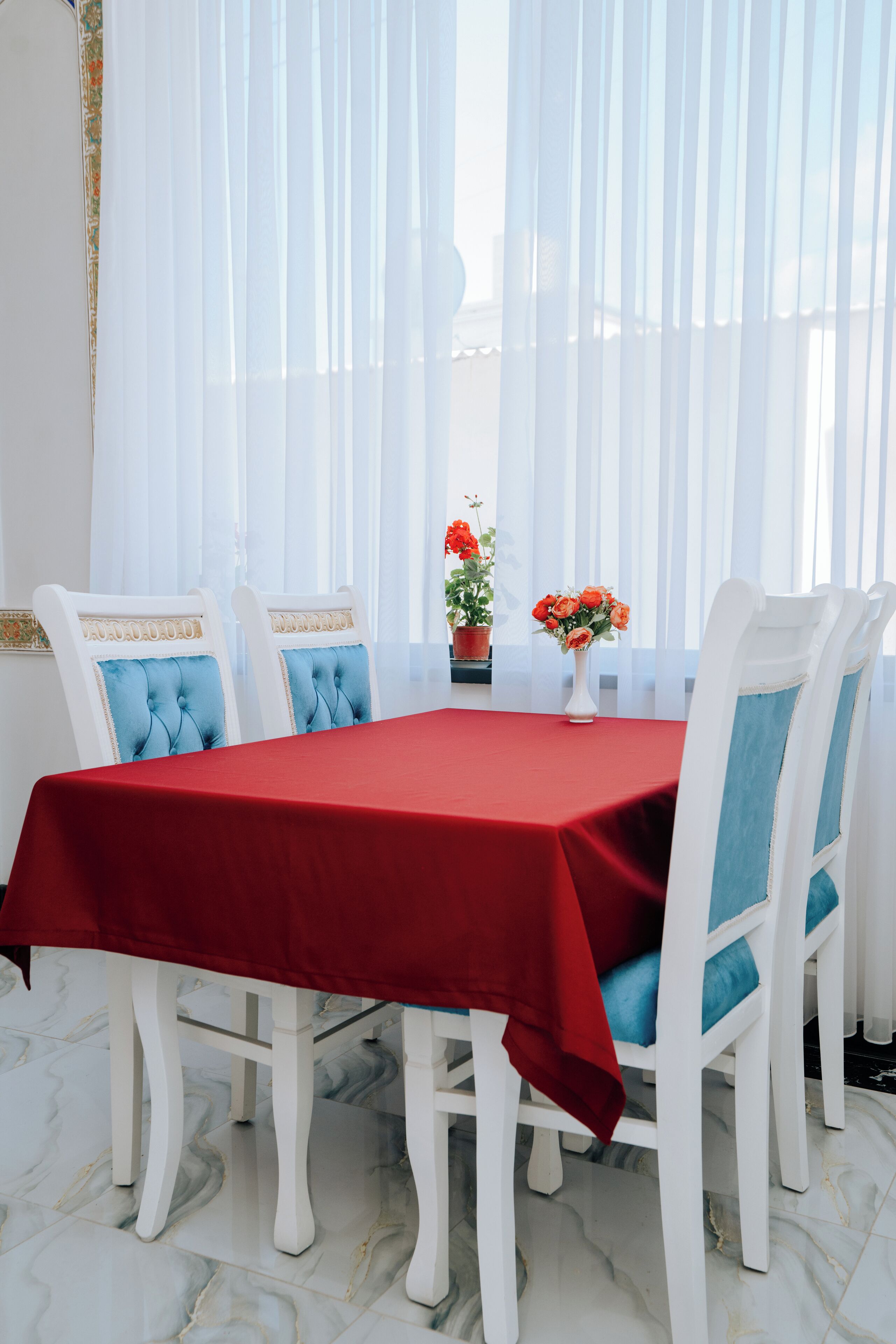 Un comedor con mantel rojo, sillas blancas con cojines azules y un florero sobre la mesa.