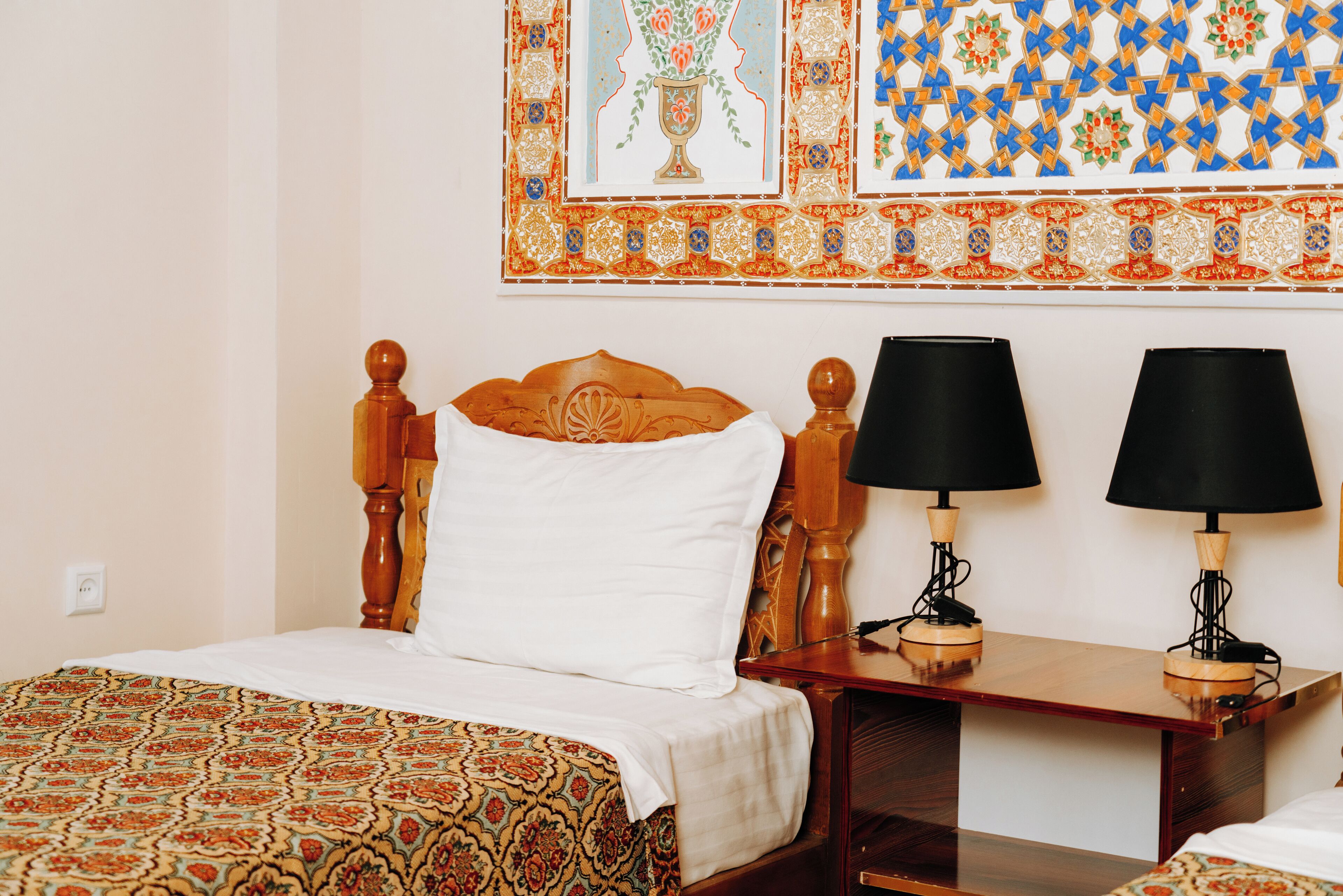 A hotel room with a wooden bed, two bedside tables with lamps, and a patterned wall hanging.