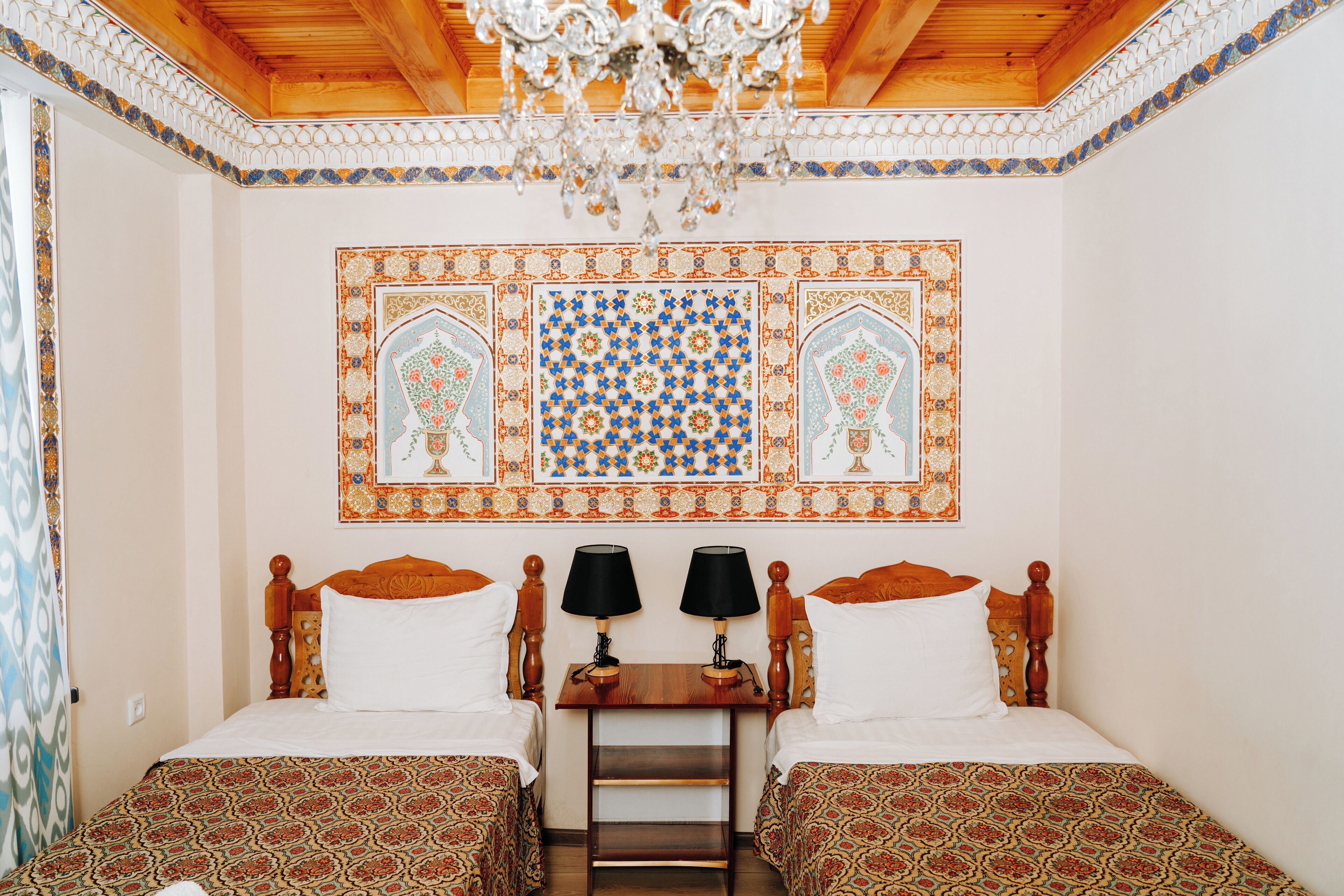 A room with two beds, a wooden headboard, a nightstand with lamps, and a decorative wall tile.