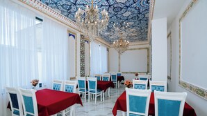 A dining area with white chairs and red tablecloths, a chandelier, and patterned ceiling. - B-House (Bukhara)