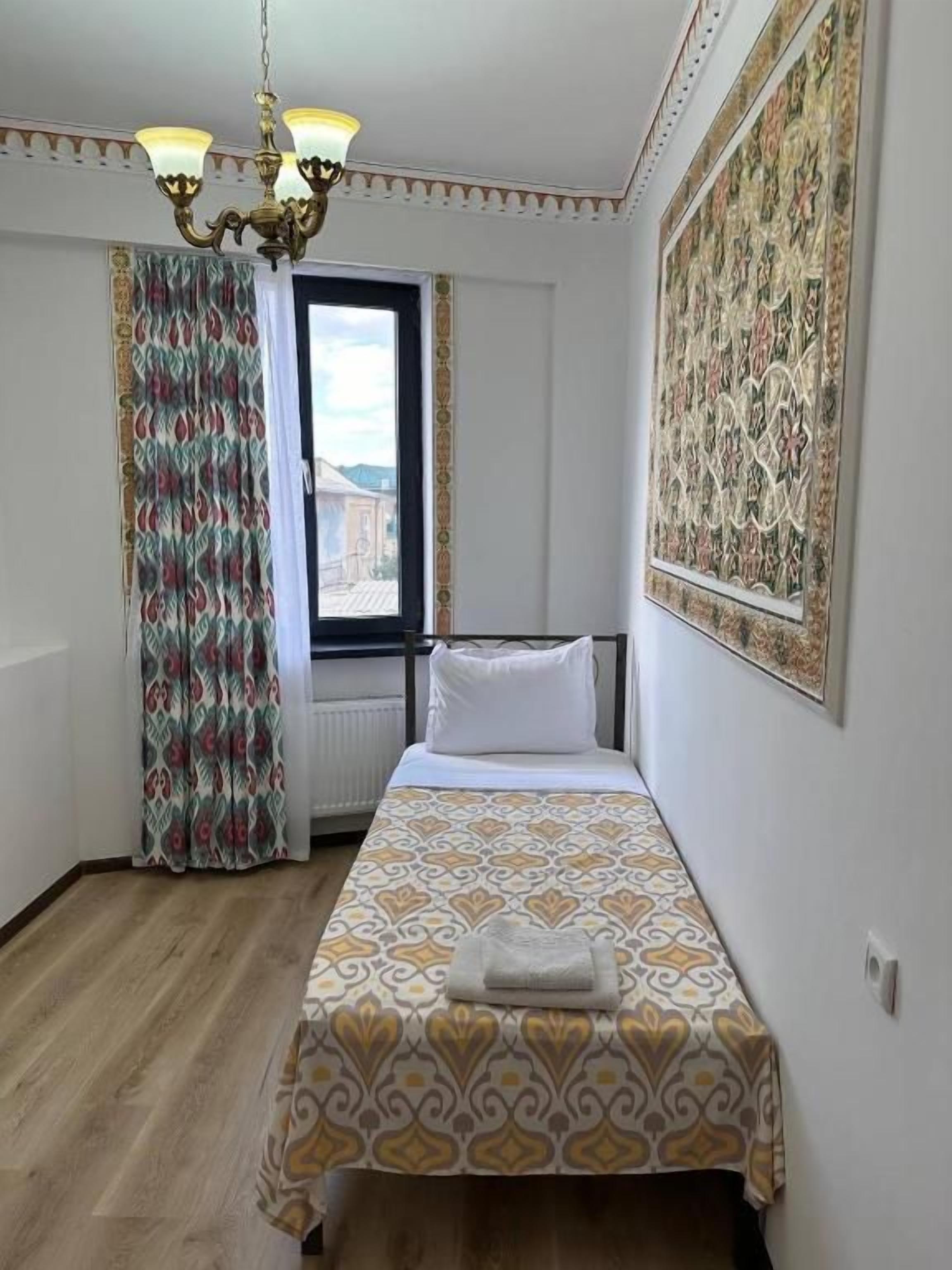 A hotel room with a bed, a window with curtains, a chandelier, and a large patterned wall decoration.