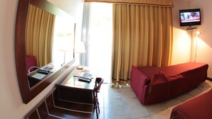 A hotel room with a sofa, a TV, a desk with a chair, and a window with curtains. - Albergo Meublé La Corte (Bagnolo San Vito)