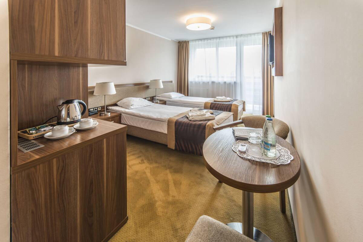 A hotel room with two single beds, a wooden desk with a kettle, a table with a bottle and glasses, and a window with curtains.
