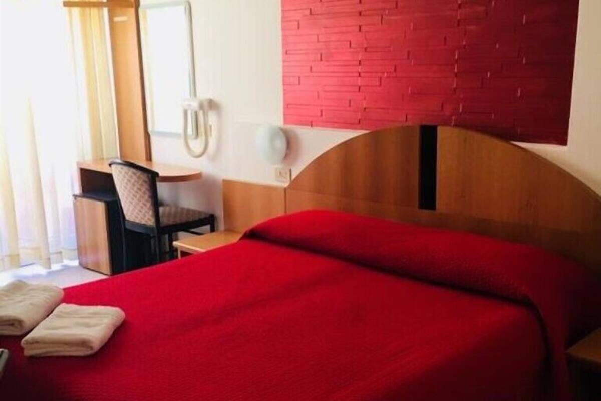 A hotel room with a red bedspread, a wooden headboard, a desk with a chair, and a wall-mounted telephone.