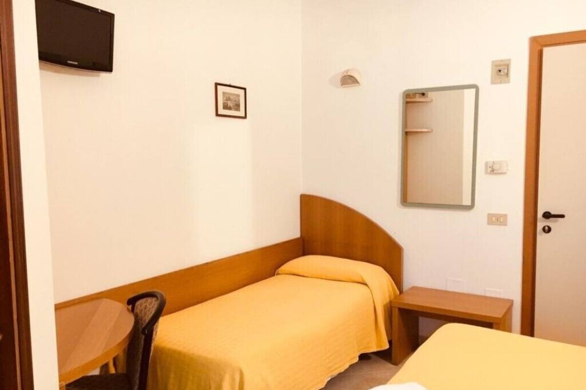 A small, single-room hotel room with a bed, a chair, a small table, a TV mounted on the wall, and a door with a handle.