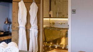 A hotel room with a bed, two white robes hanging on a wooden panel, a bathroom with a golden bathtub, and a small table with a lamp. - The World Famous Tan Hill Inn (Richmond)