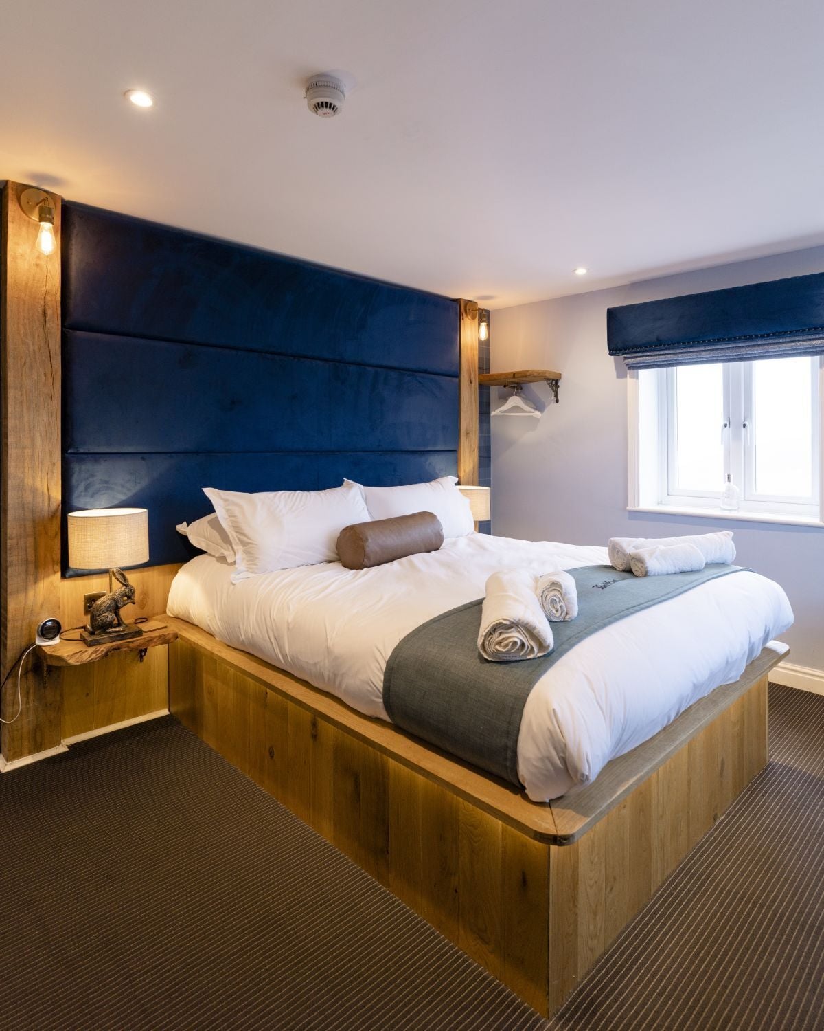 Una cama grande con estructura de madera, cabecera azul oscuro y ropa de cama blanca. Hay una mesita de noche con una lámpara y una pequeña estatua. Se ve una ventana con vistas al exterior.