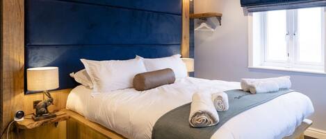 Una cama grande con estructura de madera, cabecera azul oscuro y ropa de cama blanca. Hay una mesita de noche con una lámpara y una pequeña estatua. Se ve una ventana con vistas al exterior.