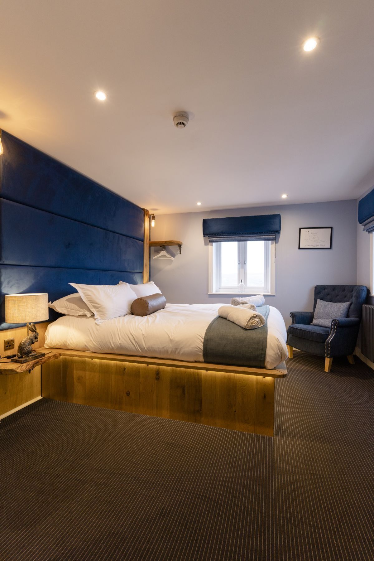 Habitación de hotel moderna con una cama grande, cabecera de madera, un escritorio con lámpara, una silla y ventana con vistas.
