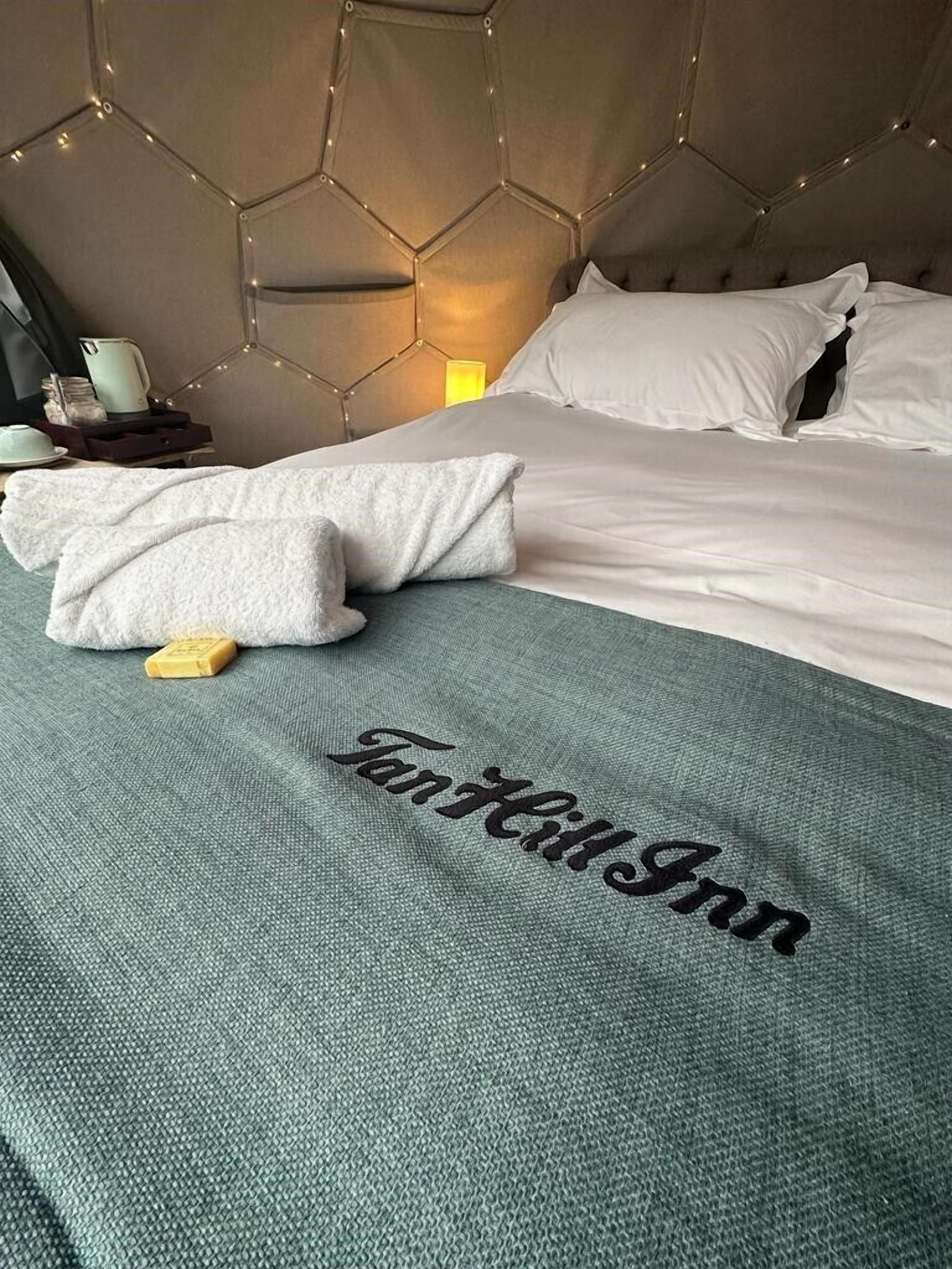 A neatly made bed with a green blanket and a towel, a bedside table with a lamp, and a hexagonal headboard with lights.