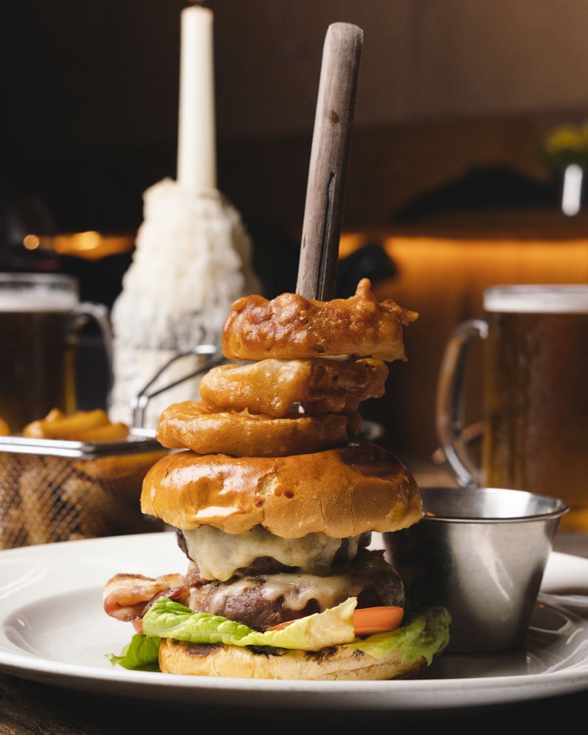 A burger with a stack of fried items on top, a metal cup, and a white mug in the background.