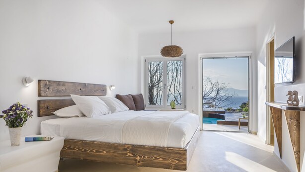 A modern bedroom with a wooden bed frame, white bedding, a bedside table with a vase of flowers, and a view of a pool and mountains through large sliding doors. - Klidon Dreamy Living Suites (Mykonos)