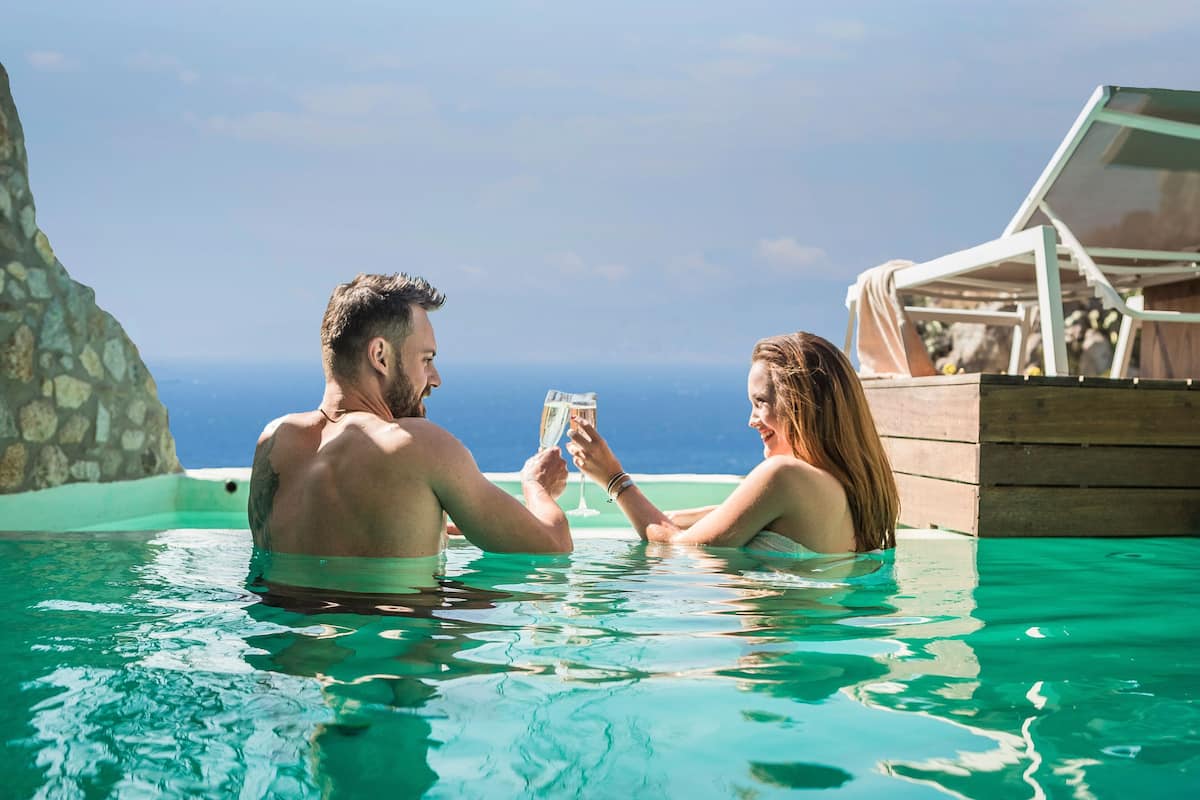 Una pareja en una piscina cristalina con terraza de madera y sillones de descanso, con vista a una extensión de agua.