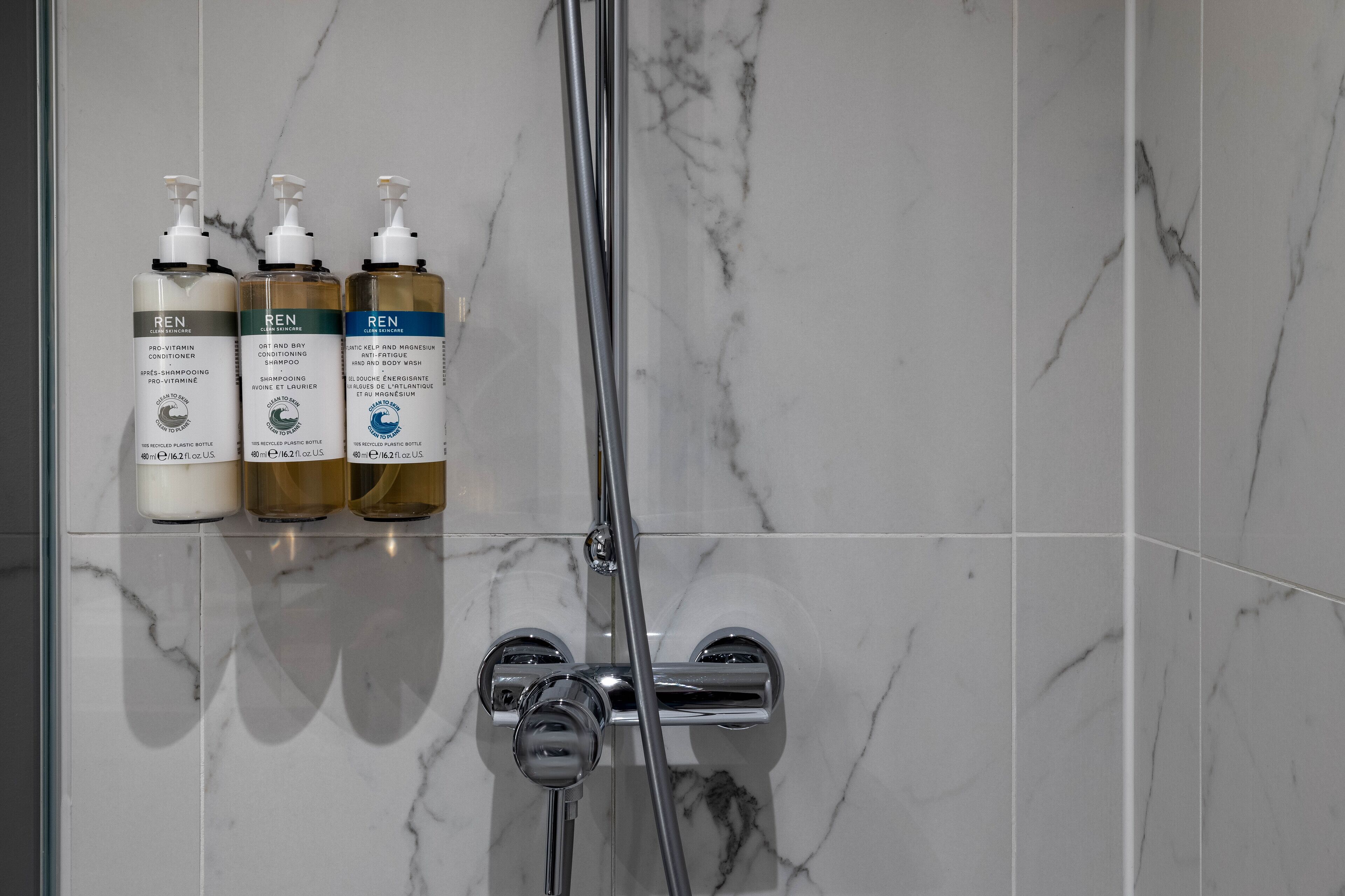 Een badkamer met een marmeren wand, een douche en drie flesjes Ren-producten.