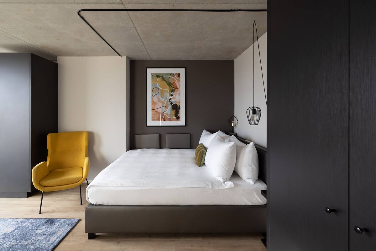 Een moderne slaapkamer met een groot bed, een gele fauteuil, een lijst met abstract schilderij en een hanglamp.