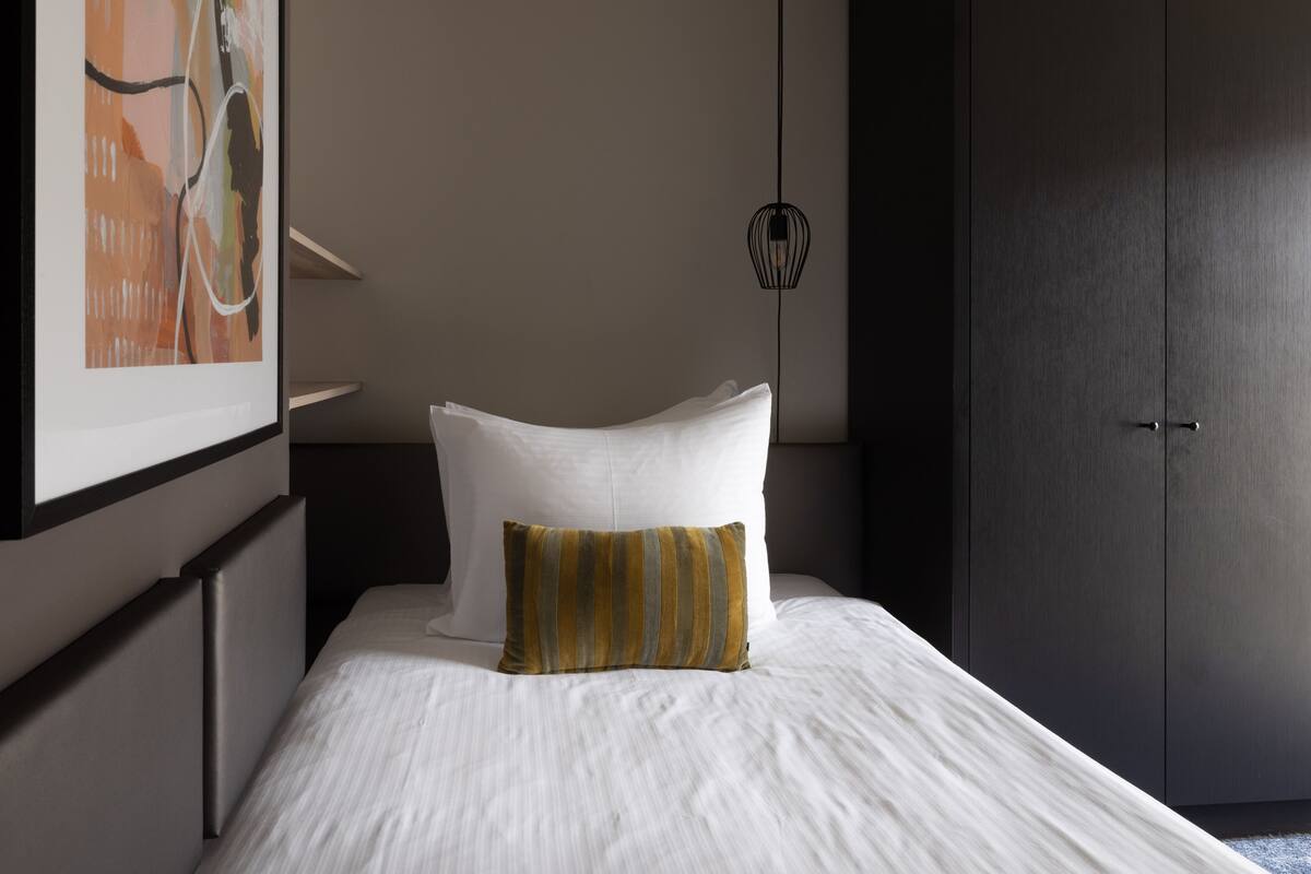 Een net opgemaakt bed met witte lakens en een gestreept kussen, een donkere kast en een wandlamp.