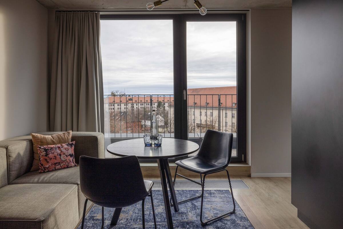 Een moderne woonkamer met een bank, twee zwarte fauteuils, een ronde tafel en een groot raam met uitzicht op gebouwen.